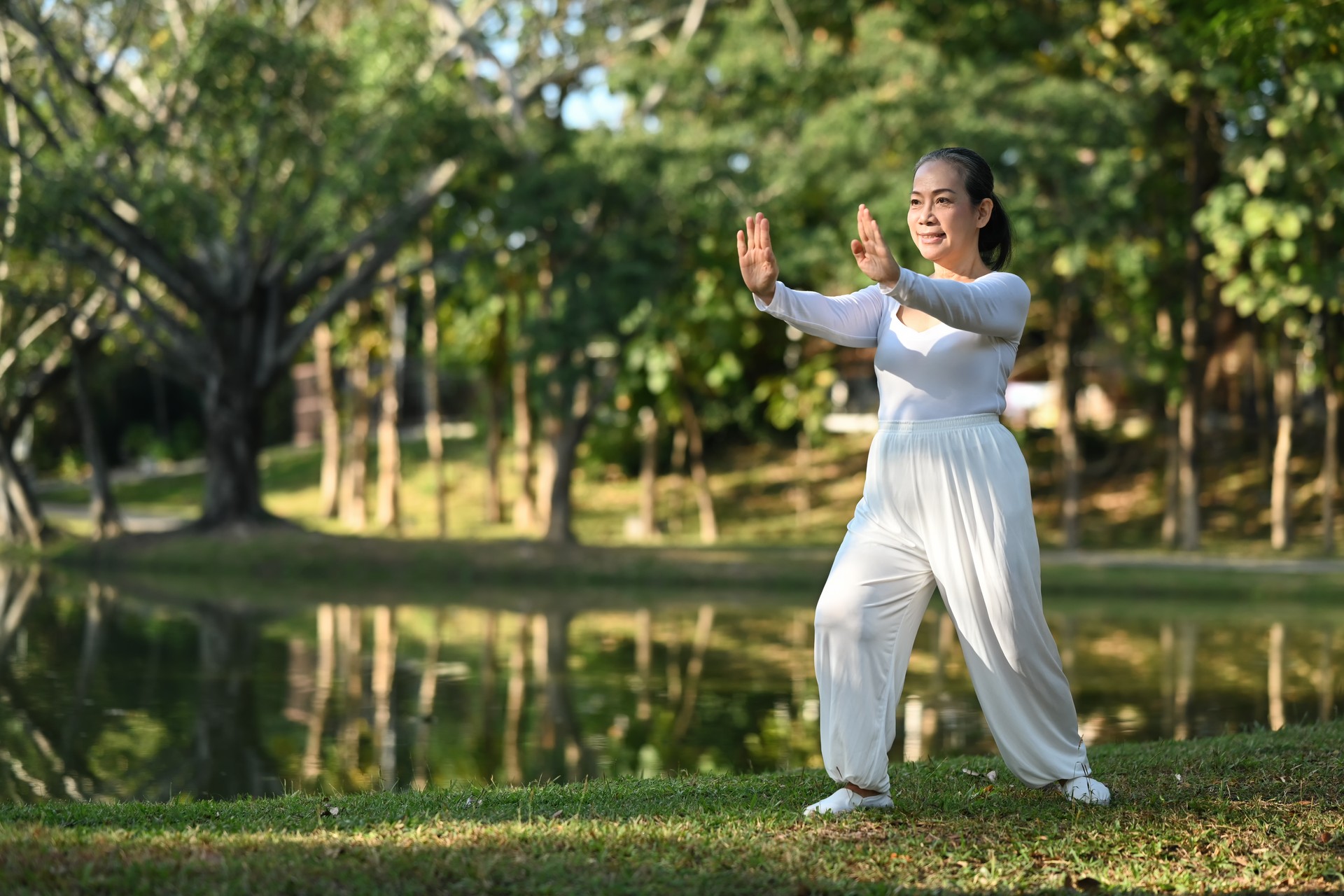 Full length of smiling senior woman practicing Qigong near lake during sunrise.