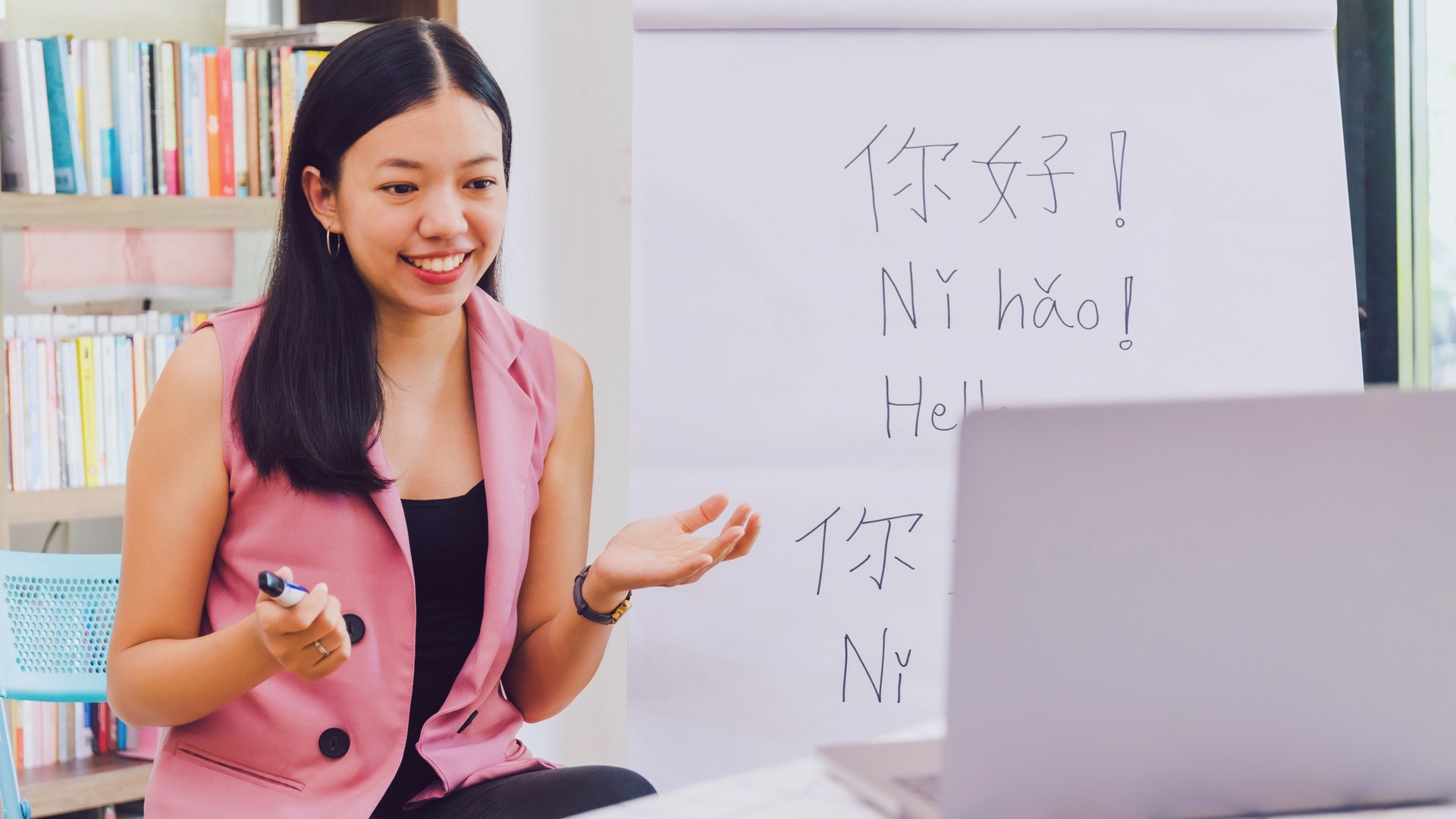 Asian woman teacher teaching remotely at home office with online technology laptop.Teaching Chinese language for Students at home school.Translation on paper text "Hello-How are you?"